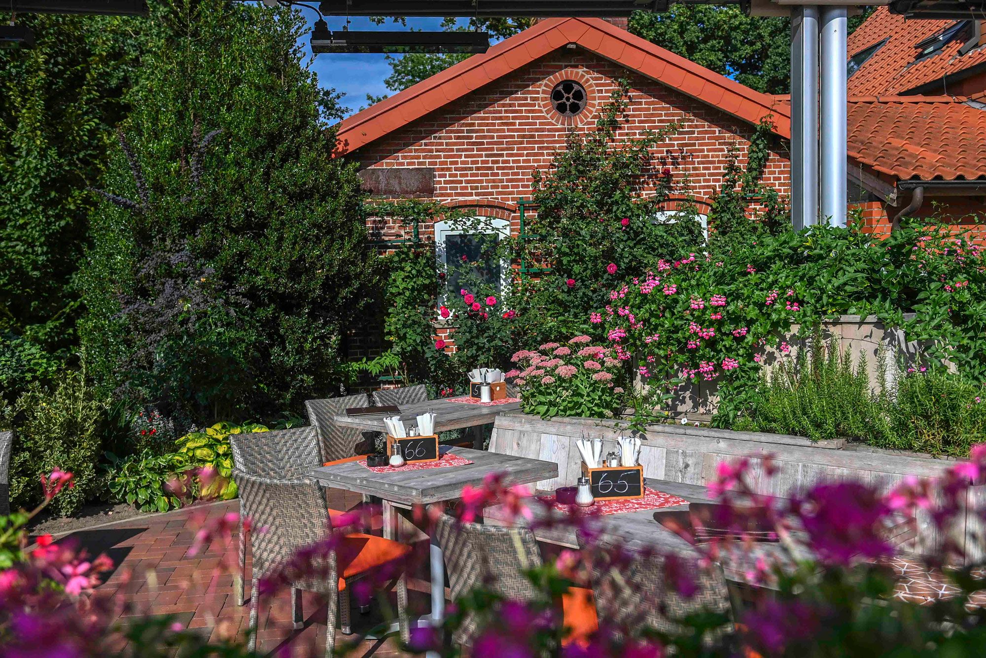 Sommergarten mit Tischen, Stühlen und Bänken. Im Vorder- und Hintergrund rosafarbene Blumen